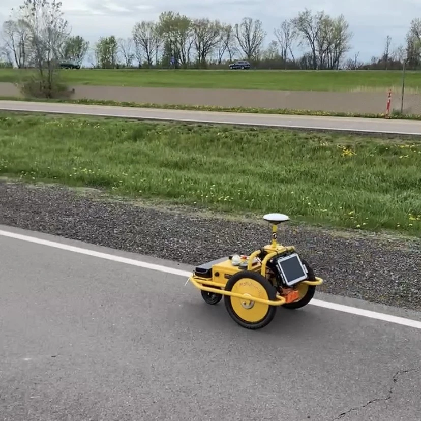 An image of a small yellow, three-wheeled remote-controlled Density Profiling System sitting on a roadway to measure the pavement's density.